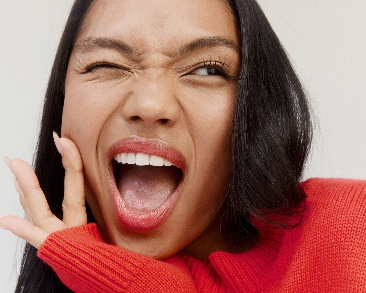 Woman in a red sweater winking and laughing with her mouth open, hand on her cheek