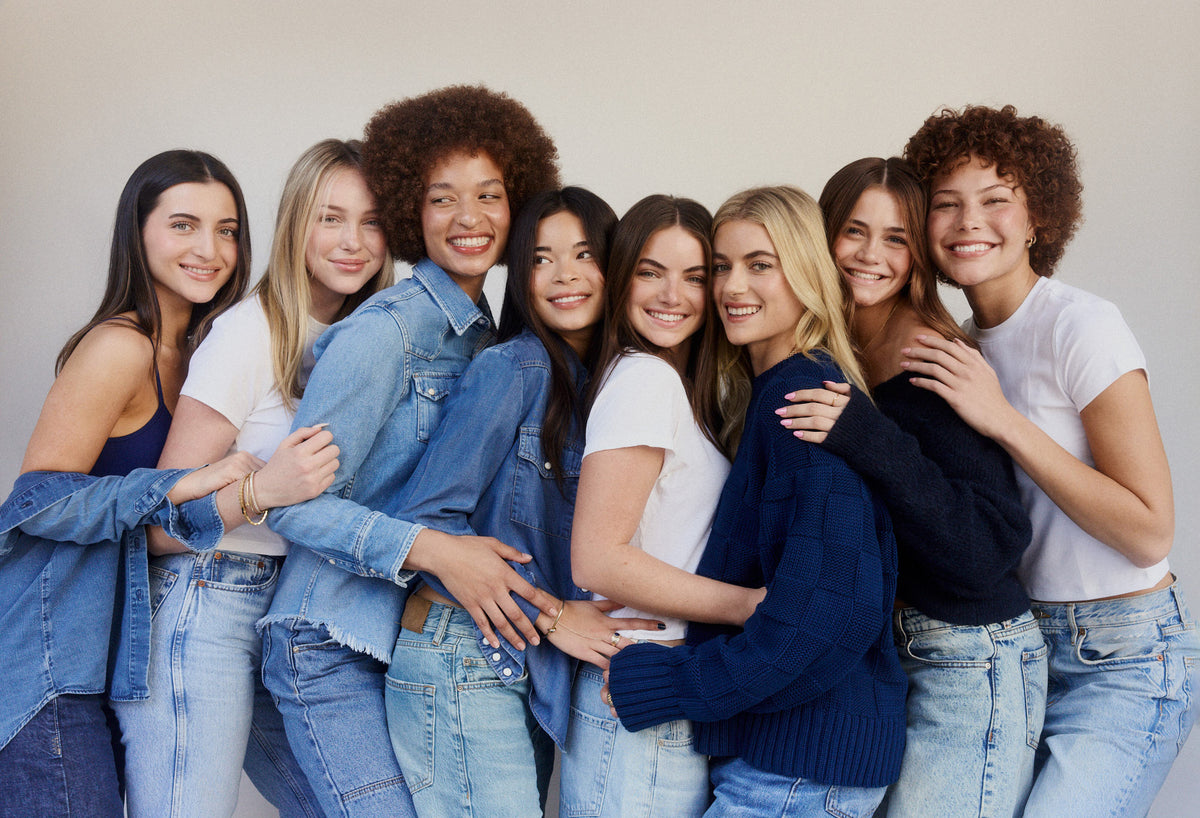 Group of eight smiling women in denim, white tees and navy knits hugging in a row against a light gray wall