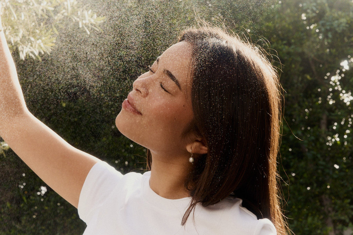 Woman in a white tee with her eyes closed and arm raised in a sunlit spray of fine mist