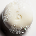 A close-up of a round white bar soap with a stylized lowercase 'b' embossed in the center, surrounded by rich lather and bubbles on a clean white background.
