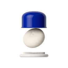 A close-up of a small personal care product with a glossy cobalt blue cap, a smooth off-white oval central piece embossed with a lowercase 'b', and a white bristle base, shown against a solid black background.