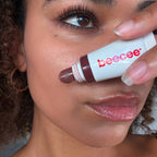 Woman holding a Jelly Gloss tube with a deep plum tip to her face, lips glossy