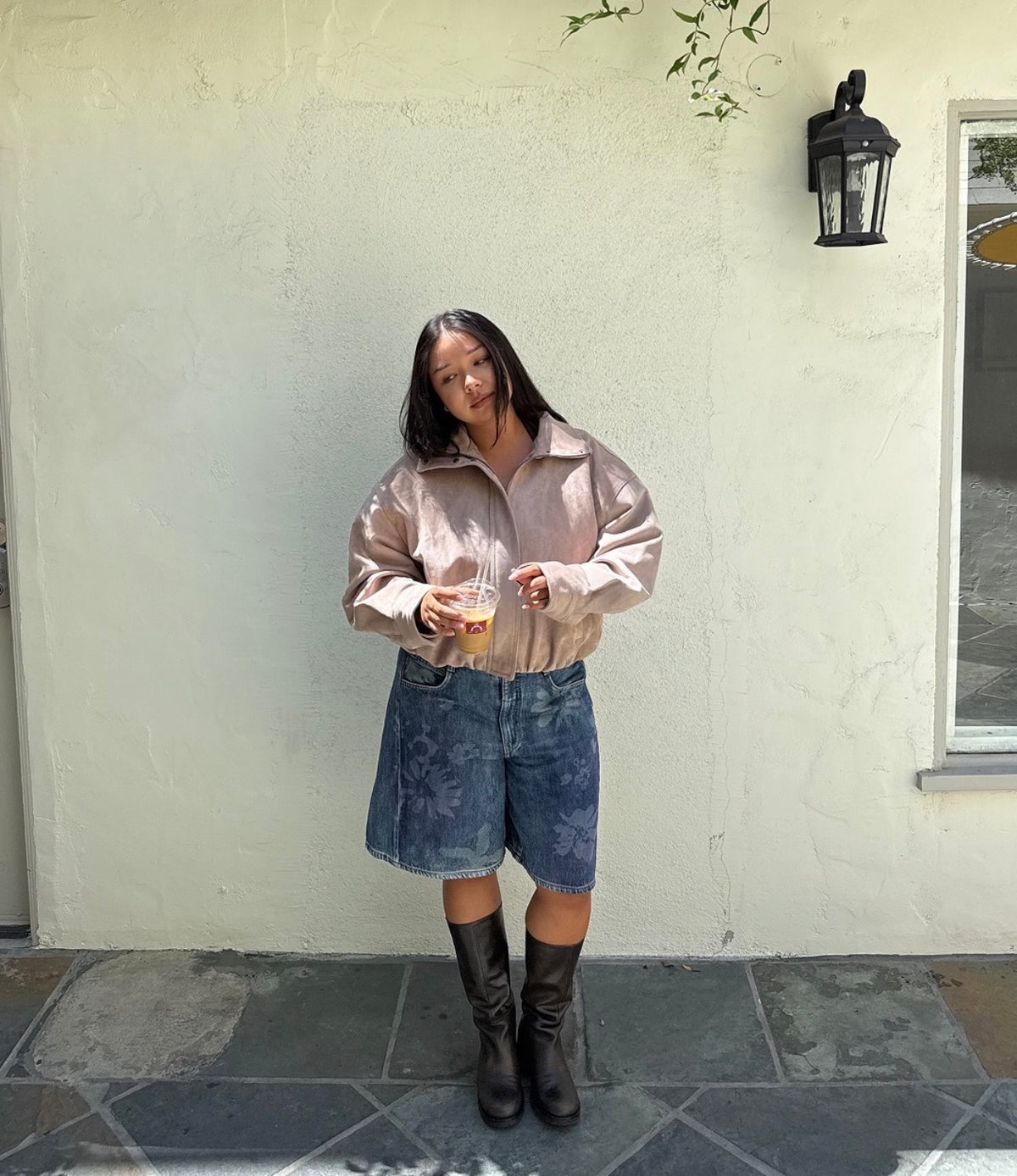 Woman in a suede jacket, denim shorts and black boots holding an iced drink against a white wall