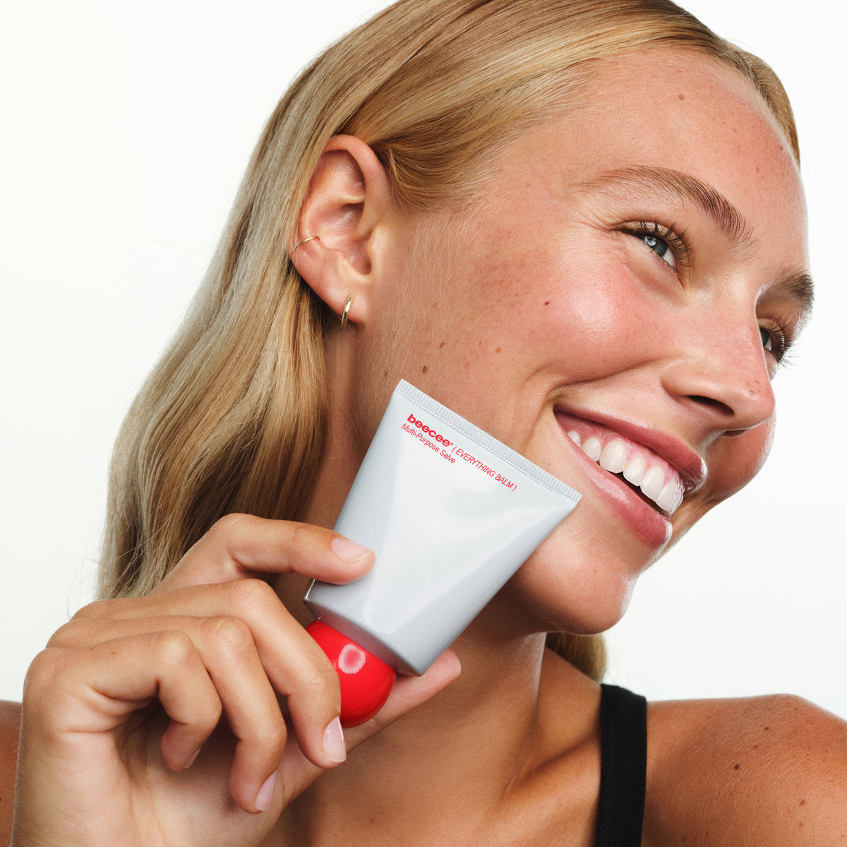 A smiling blonde woman holds a white squeeze tube of Everything balm near her face against a clean white background.