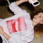 A woman lying on a concrete floor takes a selfie while wearing a white sweatshirt with a large red halftone graphic of a face on the front.