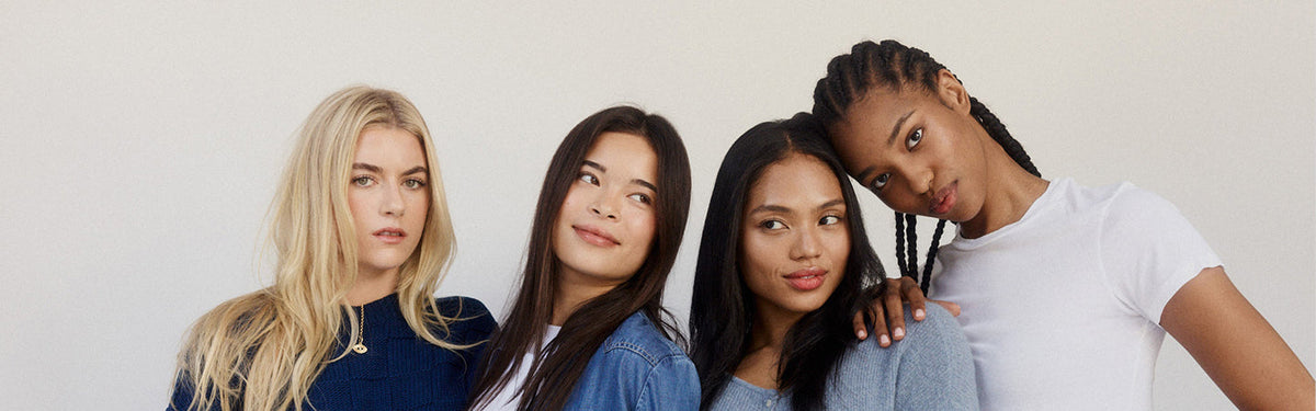 Four women posed close together against a light wall in denim and white tops