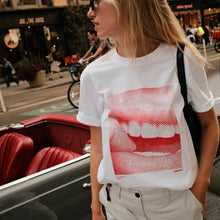 A woman wearing a white graphic t-shirt with a red halftone mouth and teeth print, standing next to a vintage red convertible car on a city street.