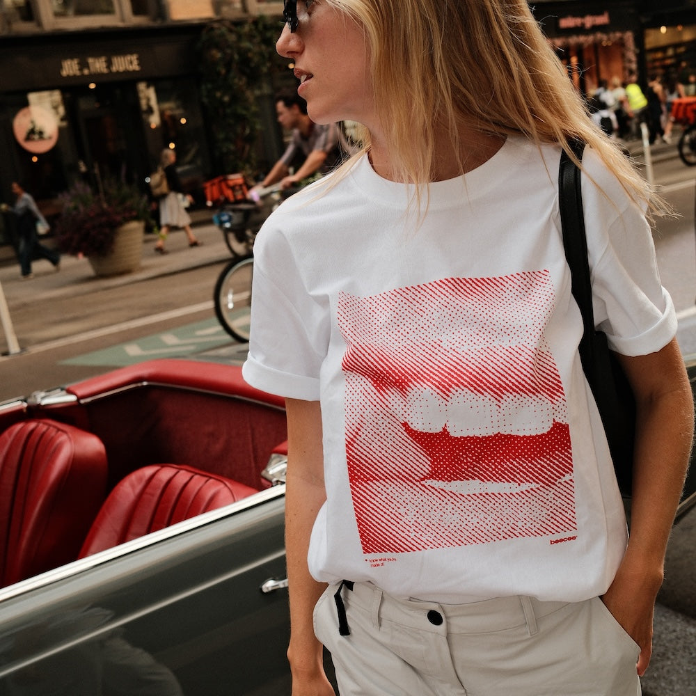 A woman wearing a white graphic t-shirt with a red halftone mouth and teeth print, standing next to a vintage red convertible car on a city street.